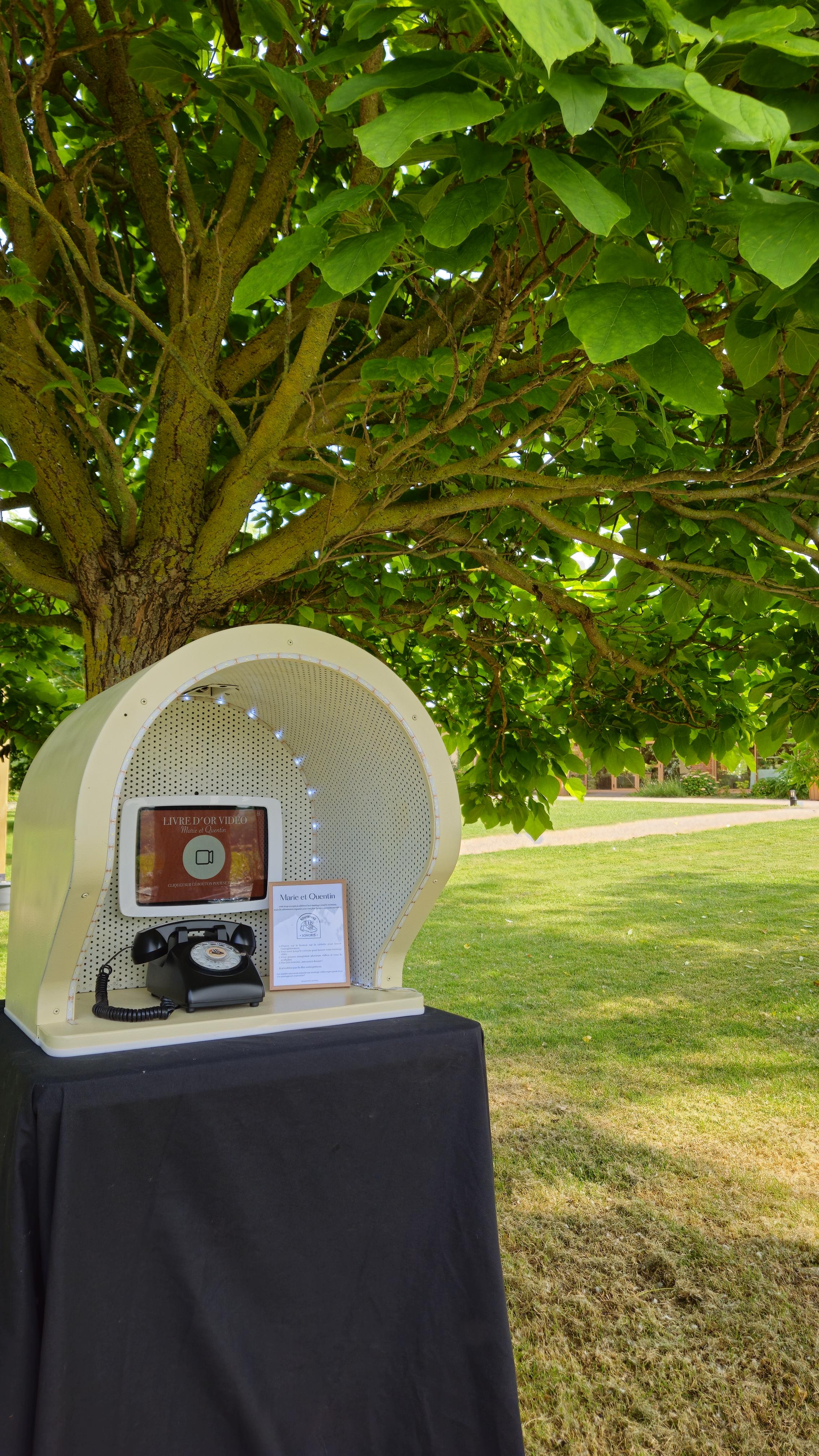 Cabine beige Souvenirs Sonores installée en extérieur sous un arbre